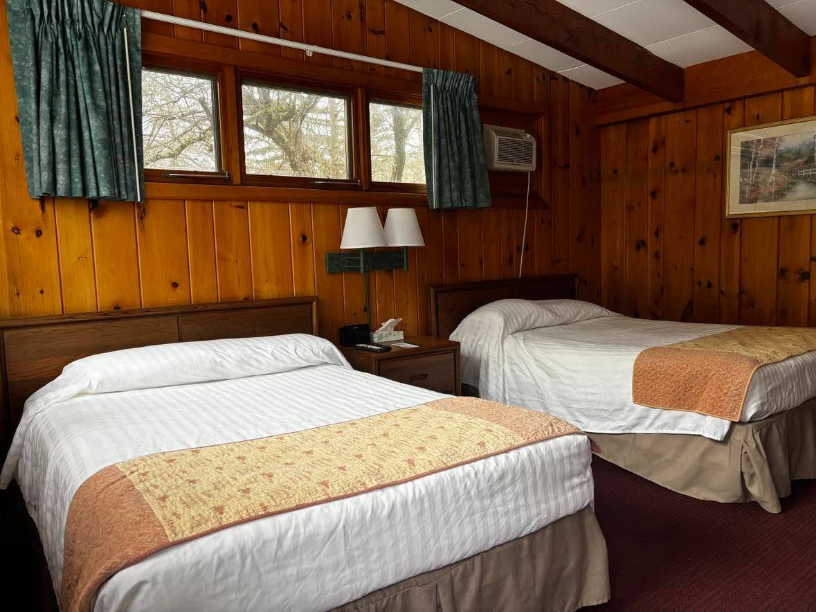 Una habitación con dos camas, paredes de madera, una ventana con cortinas, un equipo de aire acondicionado y un cuadro en la pared.