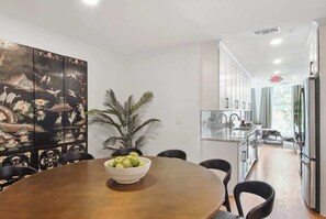 Eine moderne Küche mit Essbereich, ausgestattet mit einem großen Holztisch, einer Schüssel Äpfel, einem Blumentopf und einem dekorativen Raumteiler mit einer Naturdarstellung.
