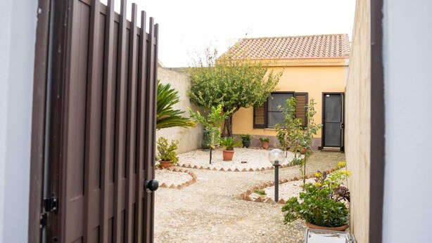 Une cour avec un chemin de gravier, des plantes en pot, une porte en bois et un bâtiment doté d’un toit en tuiles.