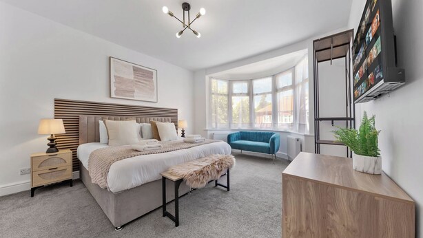 Quarto moderno com cama, mesas de cabeceira, um banquinho, sofá azul, armário de madeira, planta e uma janela grande.