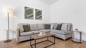 Uma sala de estar moderna com um sofá cinza em formato de L, uma mesa de centro redonda, um abajur e duas obras de arte abstratas nas paredes.