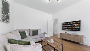 Uma sala de estar moderna com um sofá cinzento, uma mesa de centro, uma TV na parede e chão de madeira.