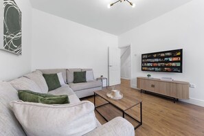 A modern living room with a grey sofa, a coffee table, a TV on the wall, and a wooden floor. - Spacious I Long stays I Sleeps 8 I Free parking (Greater London)