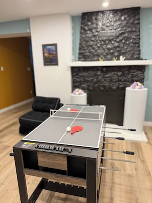 A ping pong table in a room with a stone fireplace, a framed picture on the wall, and a black sofa. - Chris Manor (Saint-Augustin-de-Desmaures)