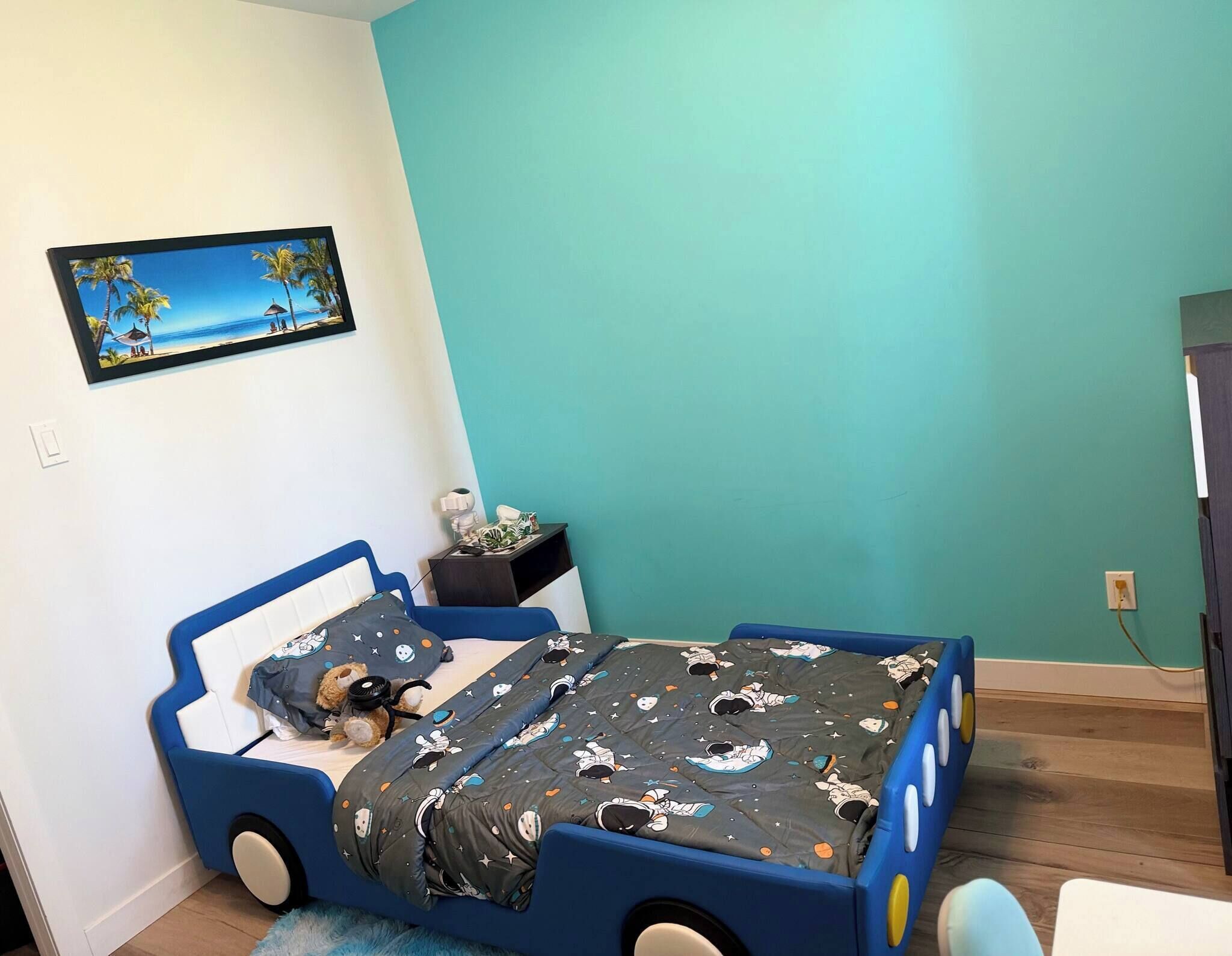 Une chambre d’enfant avec un lit, une table de chevet, un tableau accroché au mur et un plancher en bois.