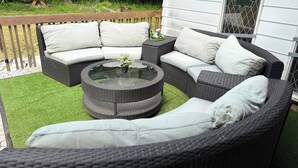 A wicker outdoor seating area with a round glass table, cushioned sofas, and a wooden deck. - Chris Manor (Saint-Augustin-de-Desmaures)
