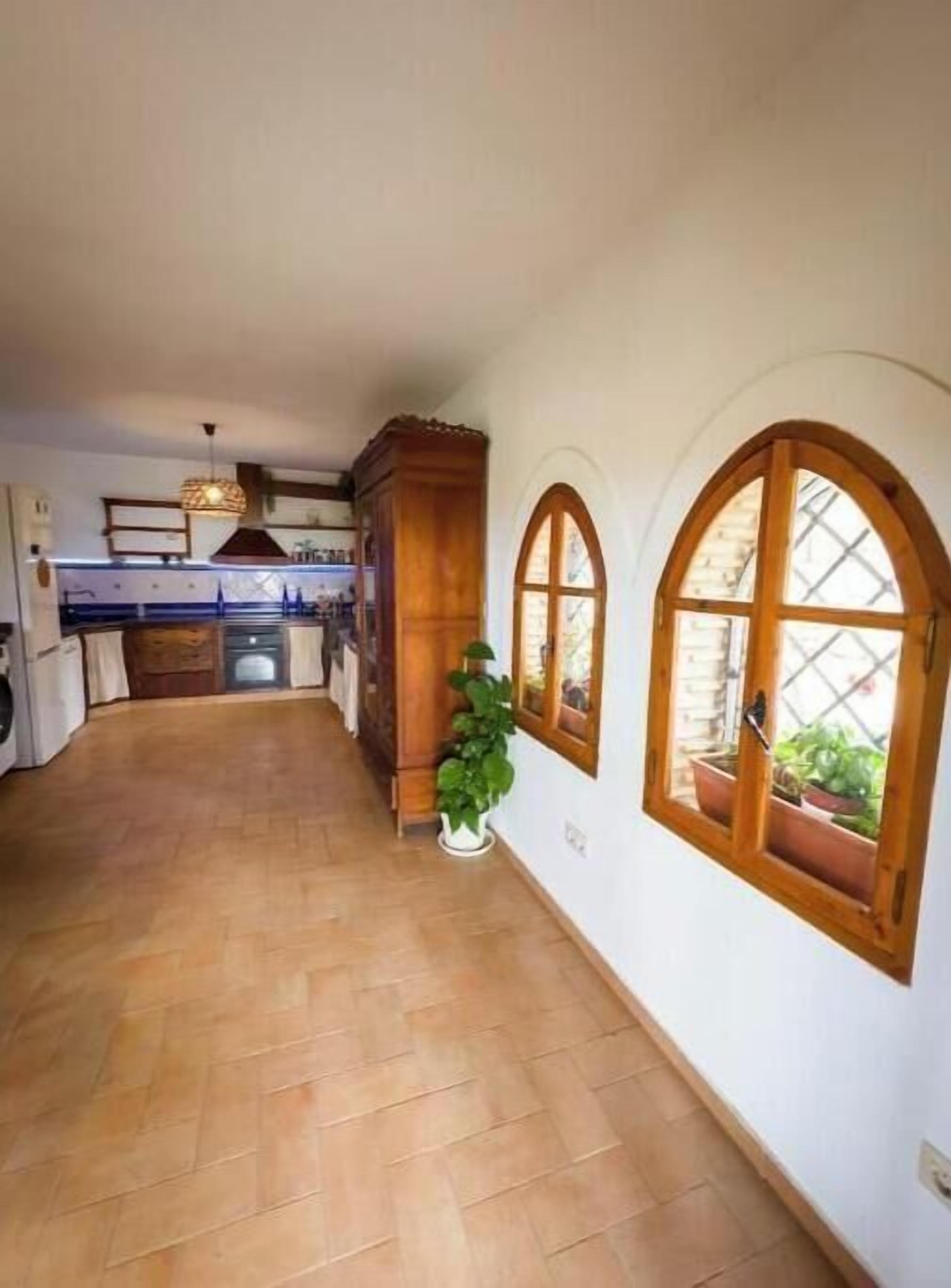 A hallway with wooden flooring, a potted plant, and two arched windows with plants.