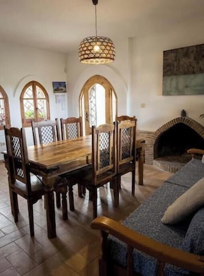 A dining area with a wooden table and chairs, a hanging light fixture, and a fireplace.