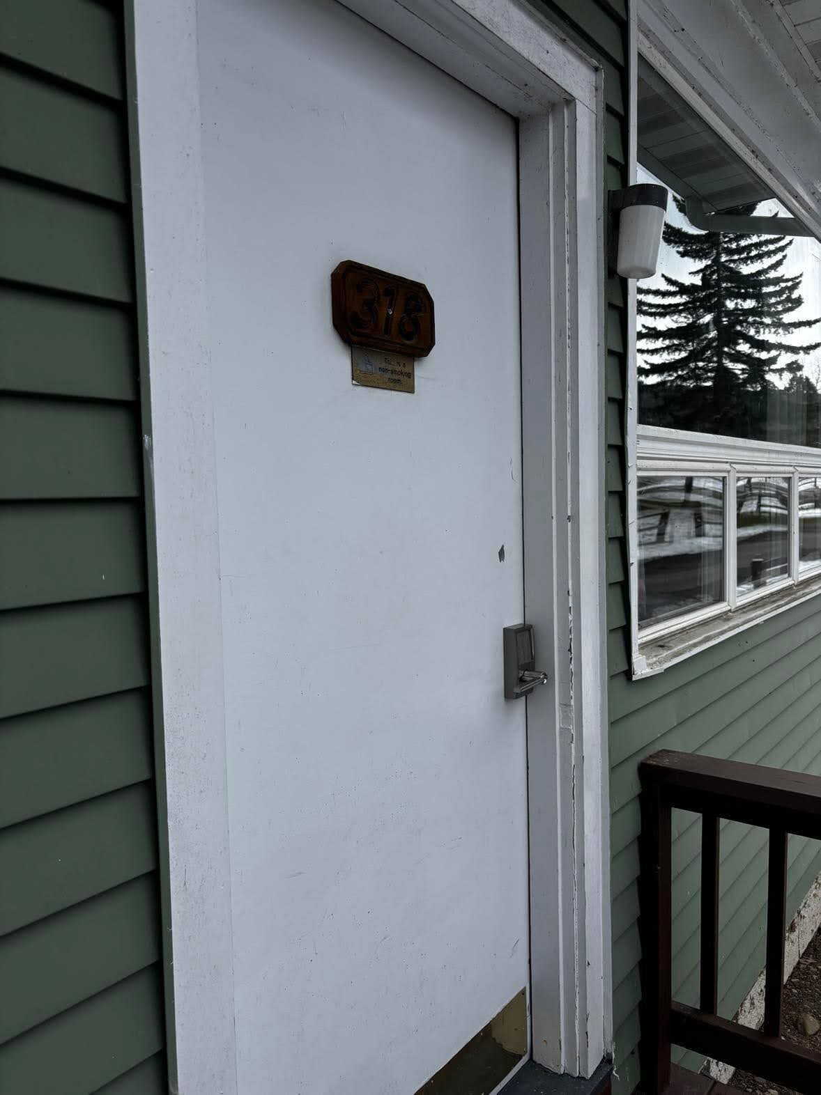 A white door with a house number plaque, a window with a view of trees, and a porch with a bench.