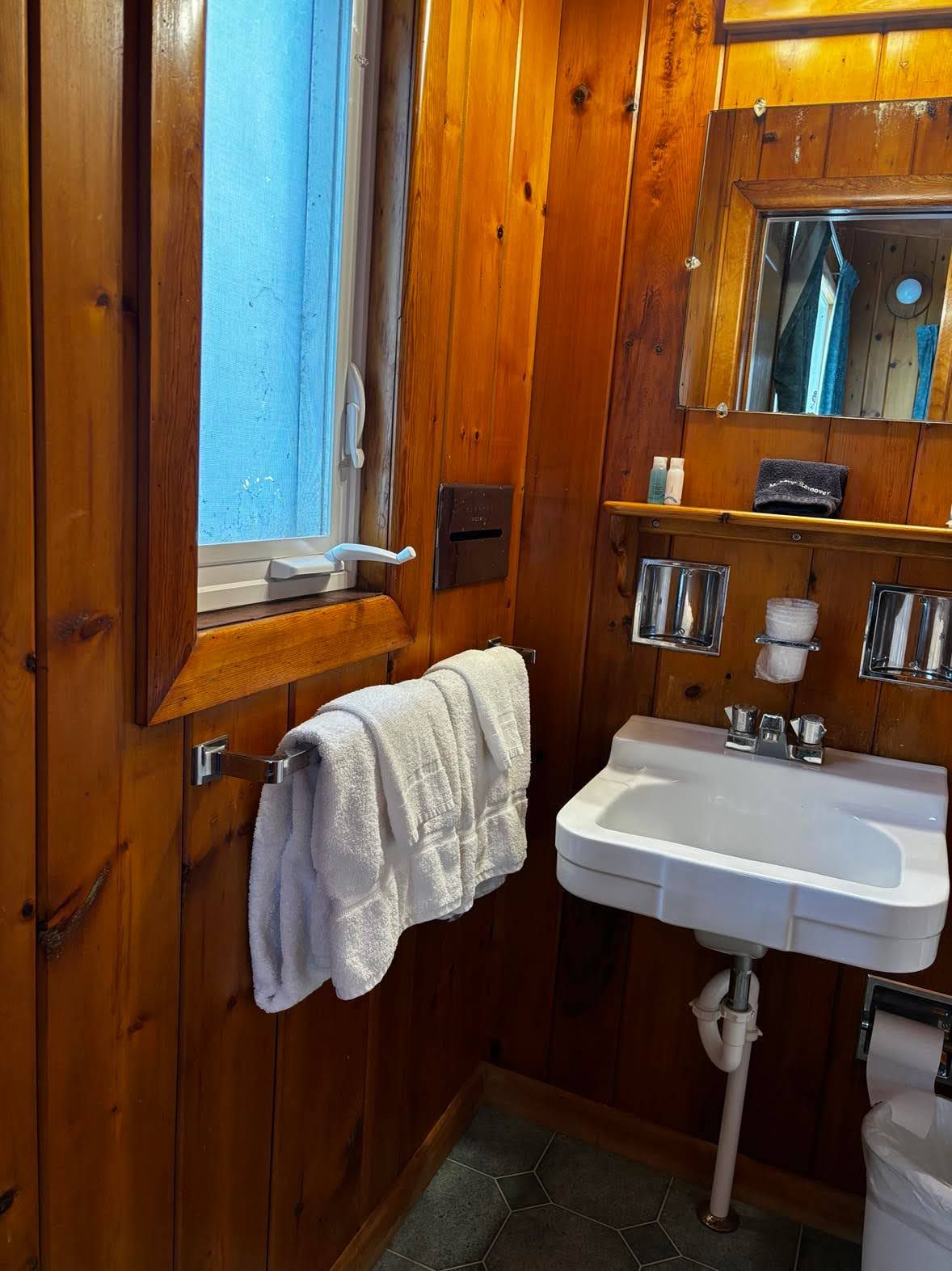 Un baño con paneles de madera, un lavamanos blanco, un perchero, un espejo y una ventana.