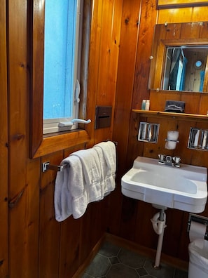 Une salle de bain aux murs lambrissés, avec un lavabo blanc, un porte-serviettes, un miroir et une fenêtre.