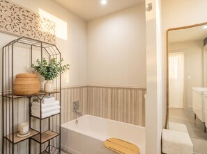 Un baño con bañera, un estante de madera con toallas y una planta, y un espejo con marco de cuerda.