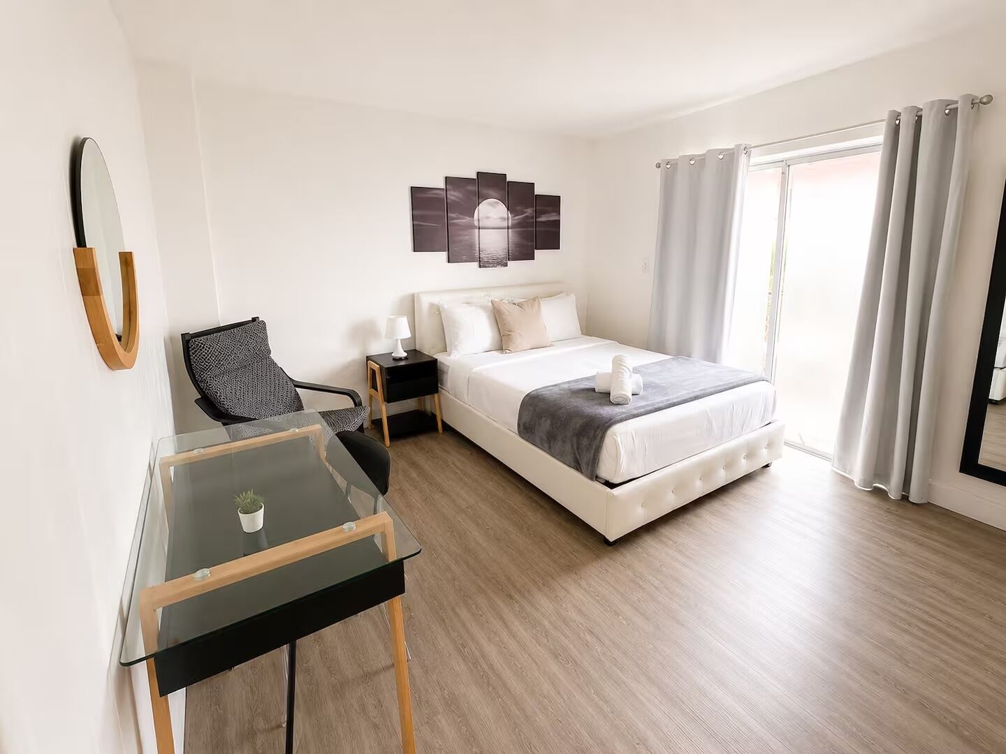 Une chambre d’hôtel comprenant un lit, un bureau, une chaise, une table de chevet, un miroir fixé au mur et une fenêtre avec des rideaux.