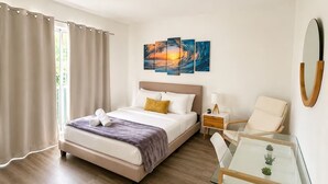 Una habitación de hotel con una cama, una mesita de noche con lámpara, una silla, una mesa de vidrio con plantas y obras de arte en la pared.