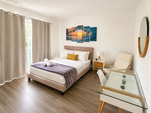 Una habitación de hotel con una cama, una mesita de noche con lámpara, una silla, una mesa de vidrio con plantas y obras de arte en la pared.