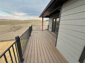 A wooden deck with a black railing, a covered area with a glass door, and a view of a hilly landscape. - Luxury Black Hills Retreat with Hot Tub, Views & Easy I-90 Access. (Box Elder)
