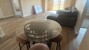Een ruime woonkamer met een eettafel met glazen blad, omringd door fauteuils, een grijze bank en een groot raam met uitzicht.