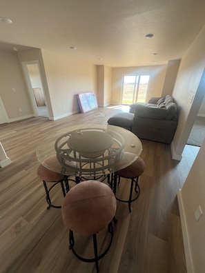 A spacious living room with a glass-top dining table surrounded by cushioned chairs, a gray sofa, and a large window offering a view. - Luxury Black Hills Retreat with Hot Tub, Views & Easy I-90 Access. (Box Elder)