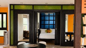 A dining area with a round table and four chairs, a bed with a blue window, and a bookshelf. - Ocean View 1BR Condo, Beach, Pool & Jacuzzi (Cabarete)