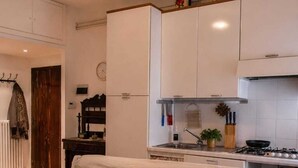 Ein gemütlicher Wohnraum mit einer Küchenzeile, einer Couch mit Kissen, einem an der Wand befestigten Kleiderhaken und einer Wanduhr.