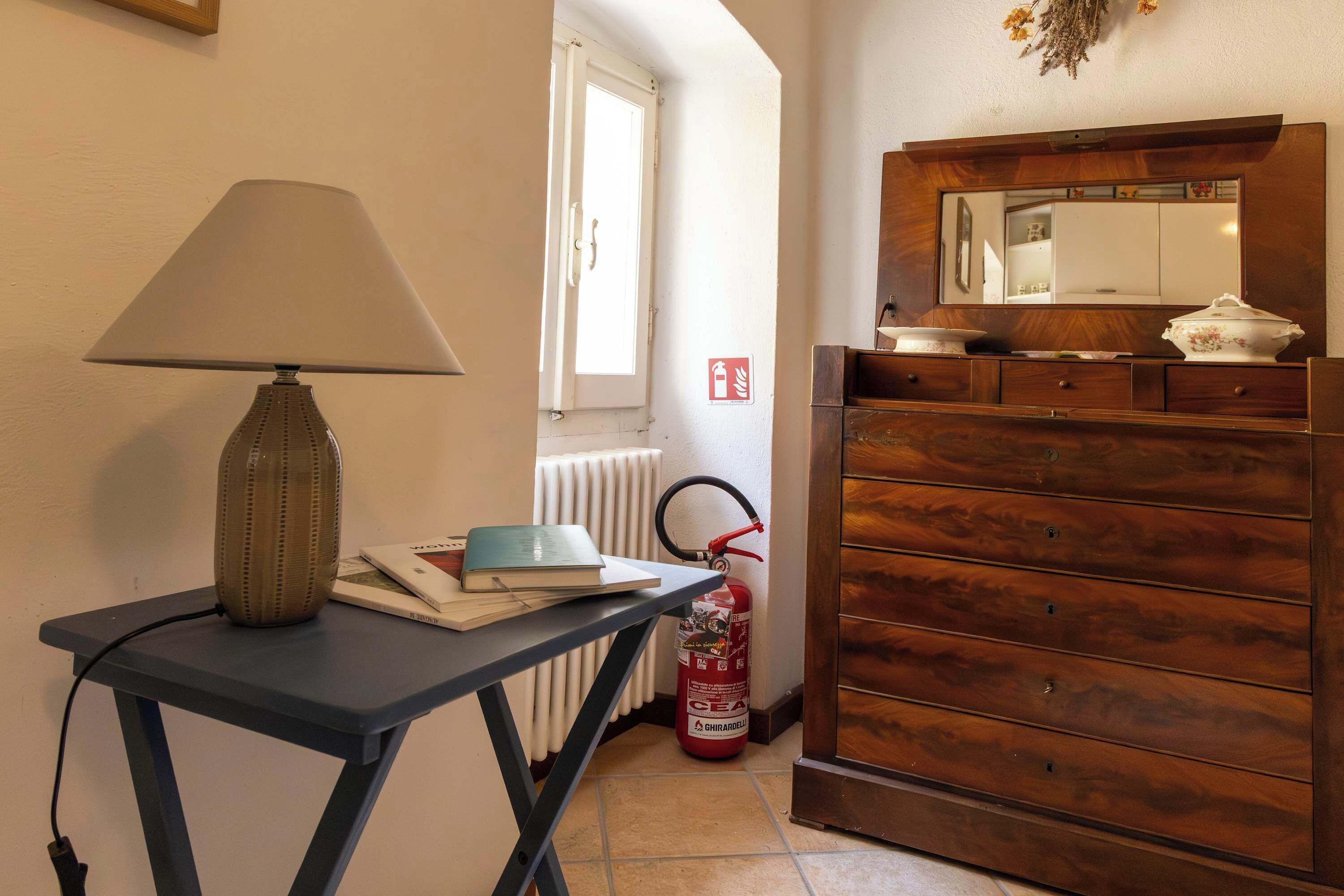 Een kamer met een tafellamp, een radiator, een brandblusser en een houten dressoir met spiegel en decoratieve objecten.