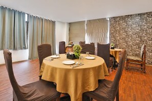 A dining area with a round table set for four, surrounded by chairs, with a stone wall and curtains in the background. - Deluxe Chandlodiya Railway (Ahmedabad)