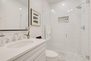 A modern bathroom with a white sink, a toilet, a shower, and a framed picture on the wall. - Luxe 8BR CBD Duplex, Private Courtyard (New Orleans)
