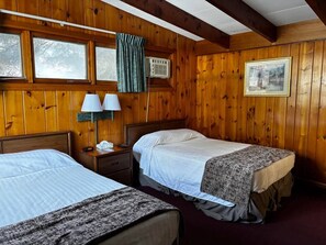 A wooden-paneled room with two beds, a nightstand with lamps, an air conditioning unit, and a framed picture on the wall.