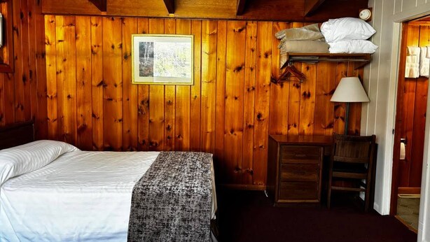 Une pièce aux murs lambrissés, avec un lit, un petit bureau, une lampe et un tableau encadré.