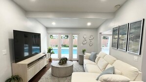 A modern living room with a flat-screen TV, a white sofa, a round coffee table, and a view of a pool through glass doors. - `Once upon a Tide`  North Palm 3BR/2B/Heated Pool (North Palm Beach)
