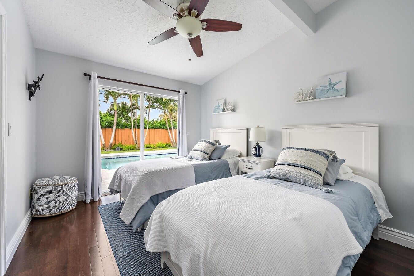 A bedroom with two beds, a ceiling fan, a window with a view of a pool, and a small side table.