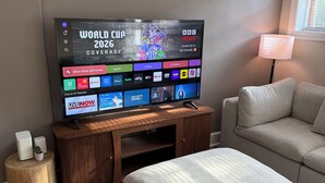 Een moderne woonkamer met een flatscreen televisie waarop de Wereldbeker 2026 te zien is, een houten tv-meubel, een grijze bank en een klein rond bijzettafeltje.