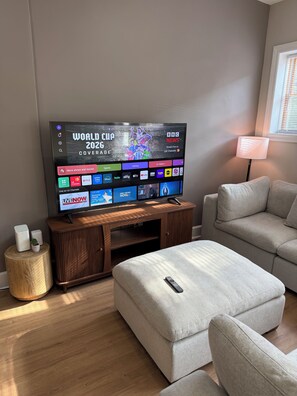 Ein modernes Wohnzimmer mit einem Flachbildfernseher, der die Weltmeisterschaft 2026 zeigt, einer Holz-Unterhaltungsecke, einem grauen Sofa und einem kleinen runden Beistelltisch.