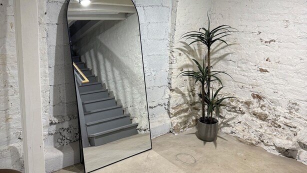 Een kamer met een grote spiegel die een trap, een potplant en een betonnen vloer weerspiegelt.