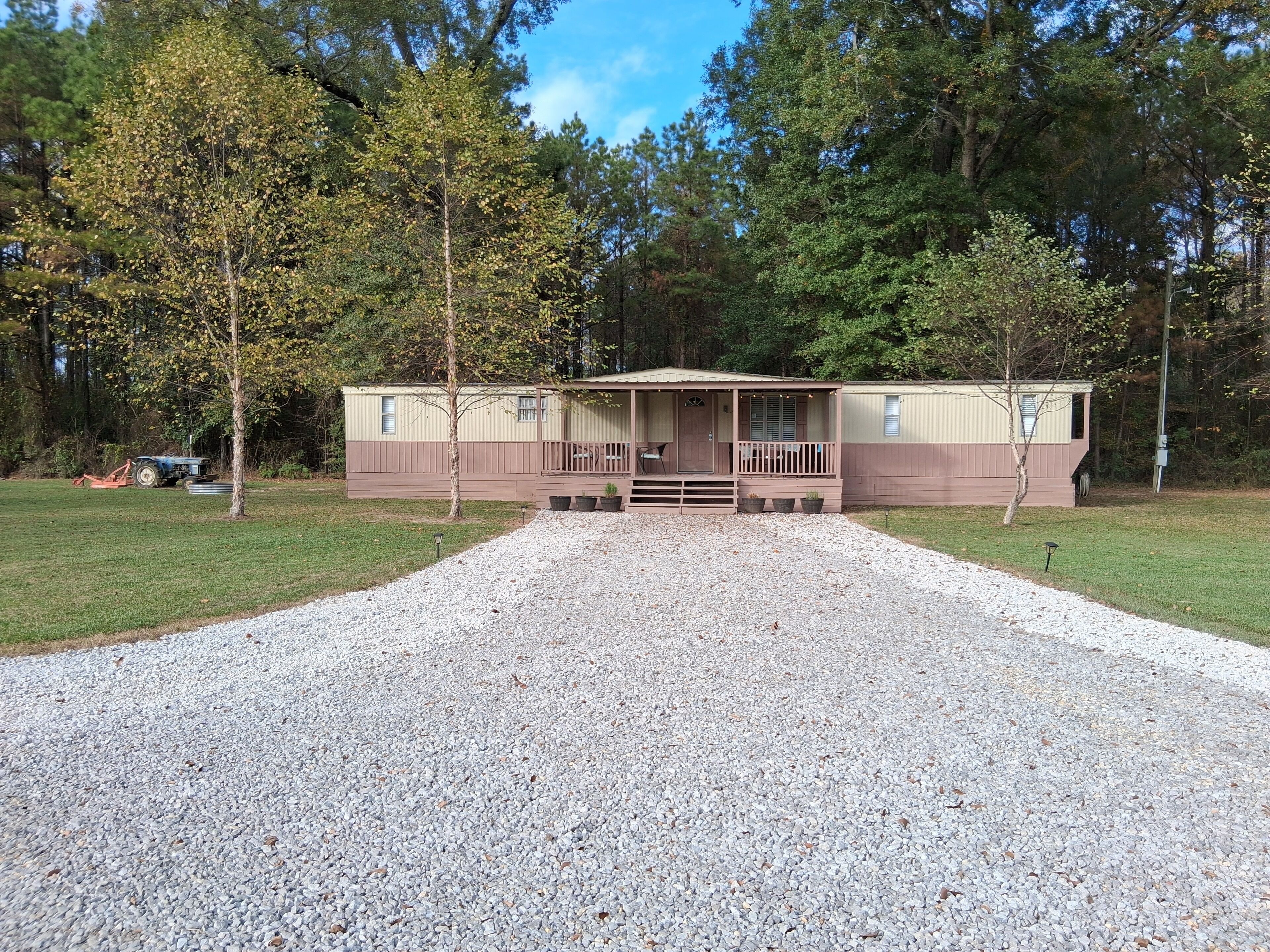 A mobile home with a porch, gravel driveway, and trees in the background.