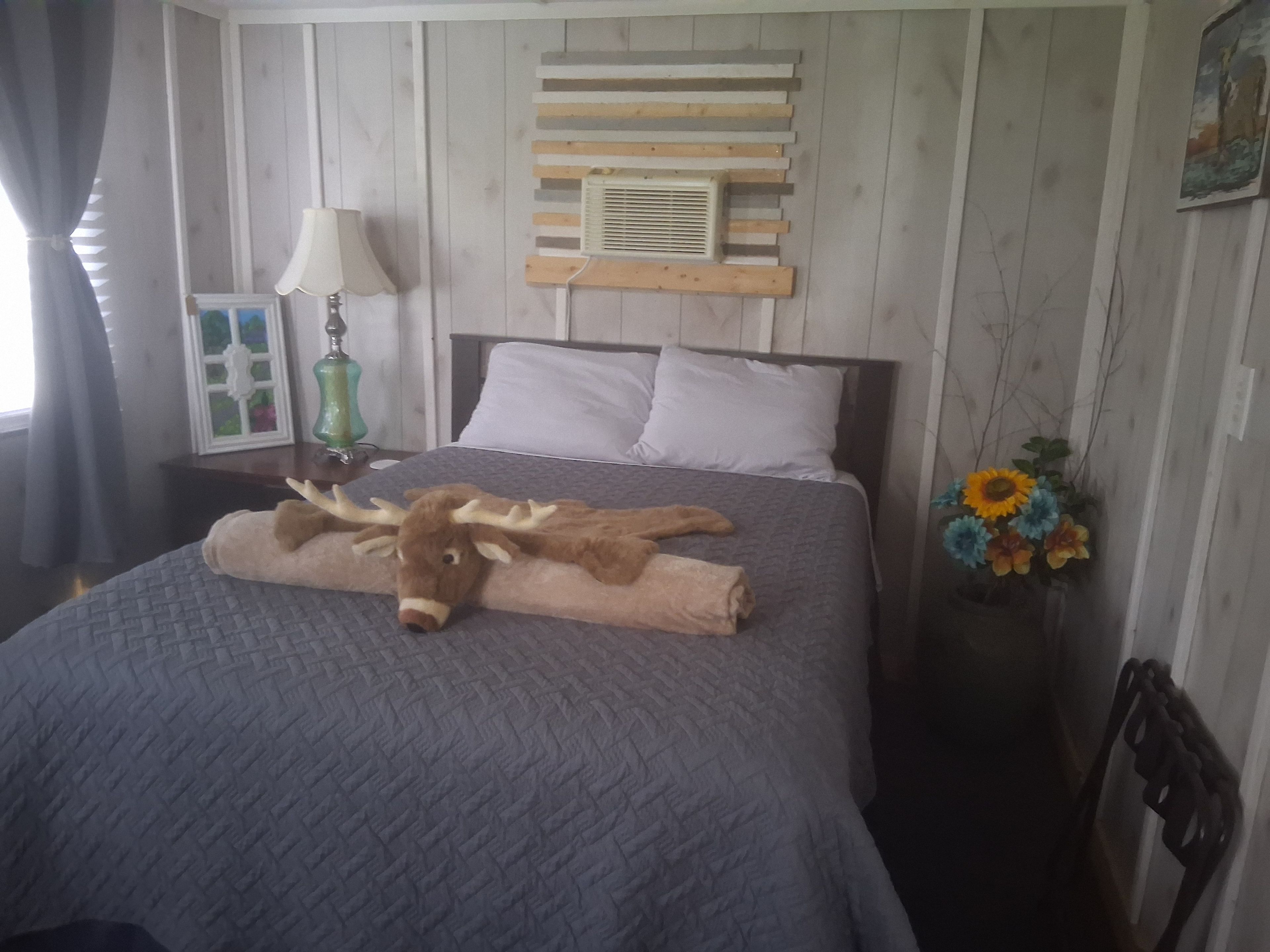 A bed with a patterned bedspread, a decorative headboard, a wall-mounted air vent, and a vase with flowers.