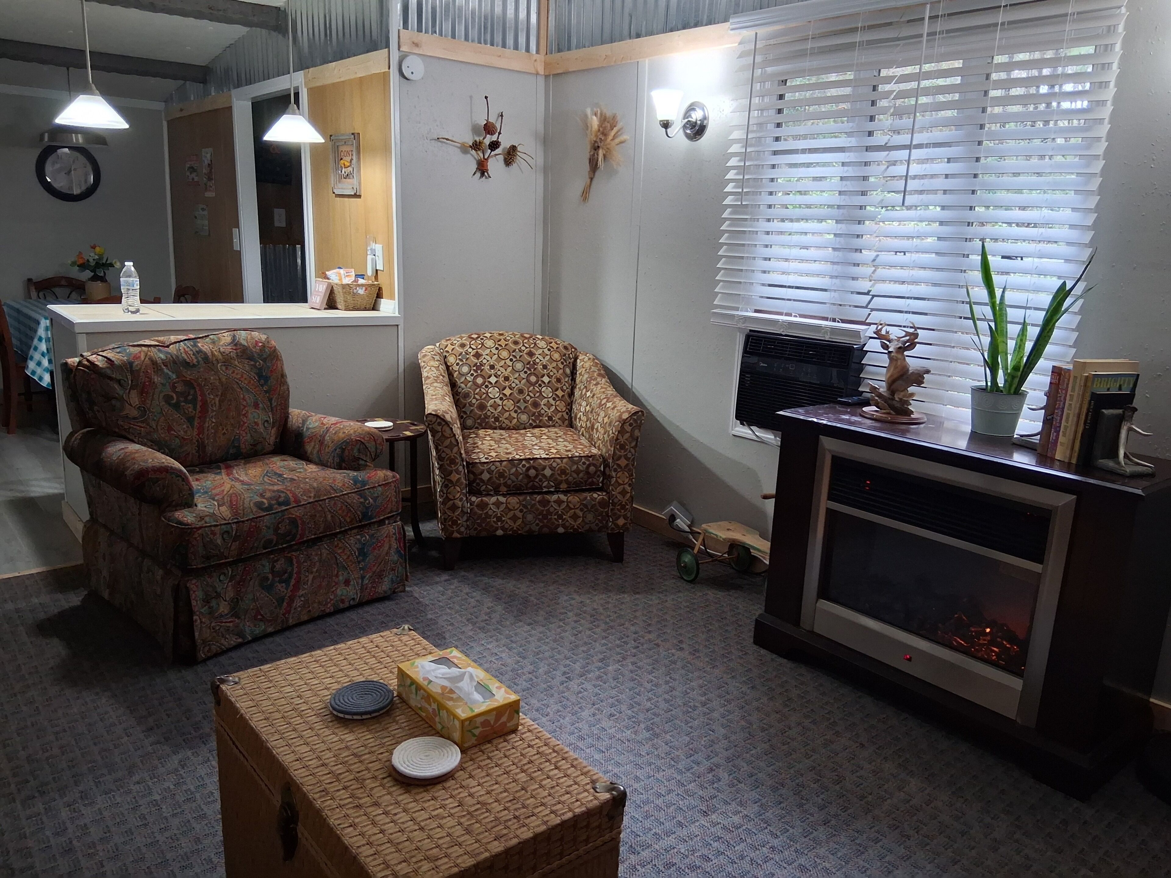 A living room with two patterned armchairs, a small table with a box and a plate, a fireplace, and a window with blinds.