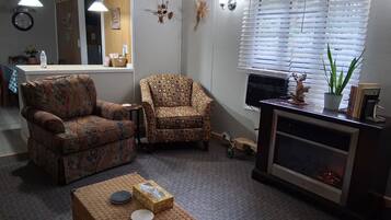 A living room with two patterned armchairs, a small table with a box and a plate, a fireplace, and a window with blinds.