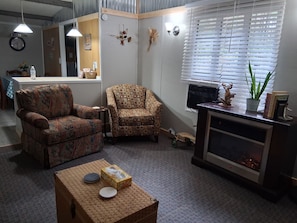 A living room with two patterned armchairs, a small table with a box and a plate, a fireplace, and a window with blinds.