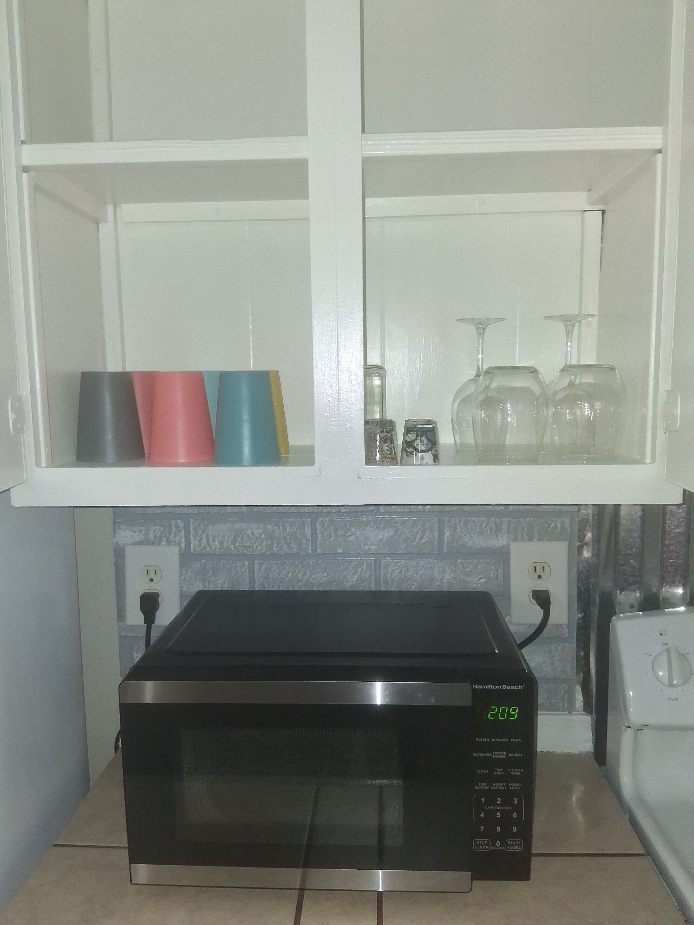 A kitchen with a black microwave, a white toaster, and a shelf with colored cups and wine glasses.