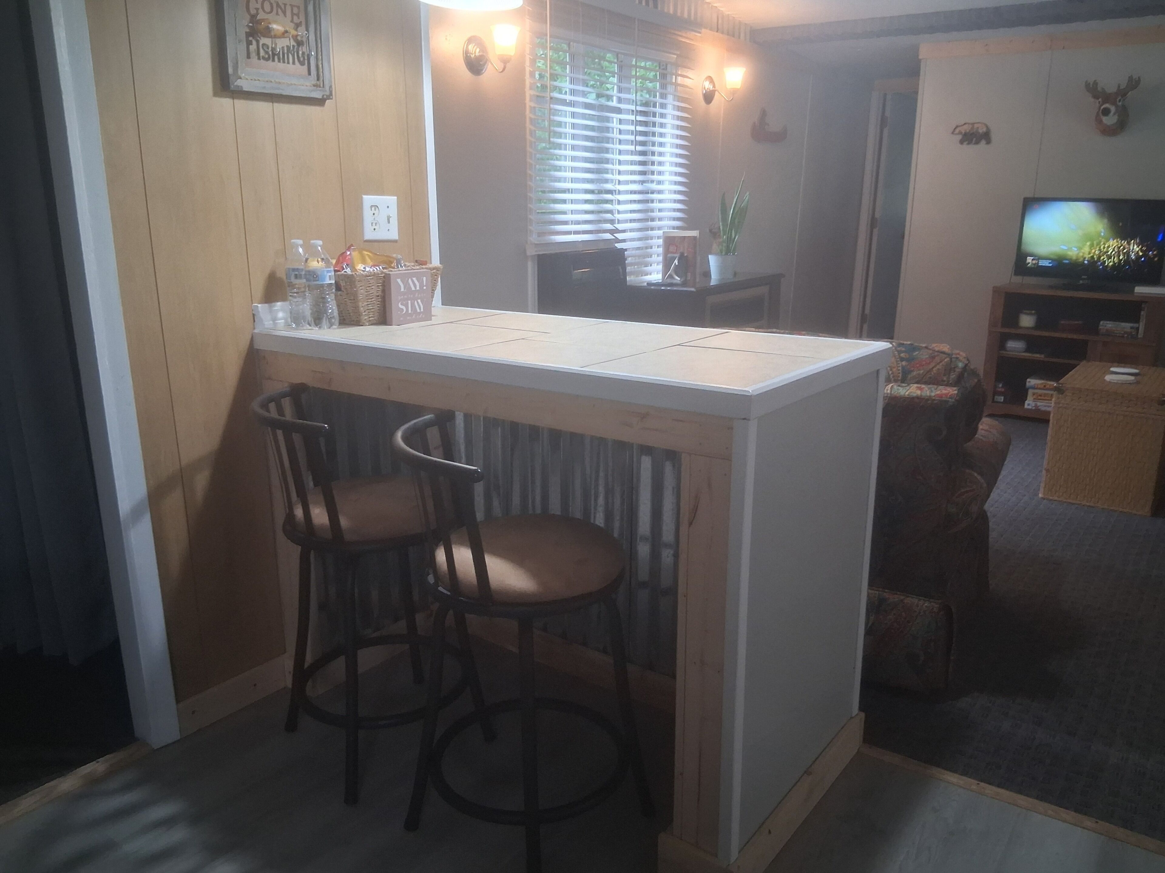 A kitchen area with a bar counter, two bar stools, a television, and a window with blinds.