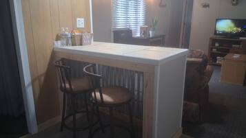 A kitchen area with a bar counter, two bar stools, a television, and a window with blinds.