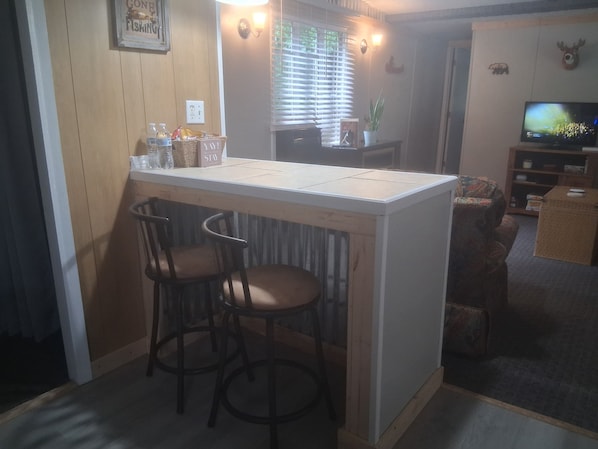 A kitchen area with a bar counter, two bar stools, a television, and a window with blinds.