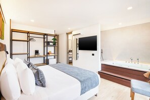 Un dormitorio moderno con cama, mesitas de noche, televisión, ducha integrada y un pequeño rincón de descanso.