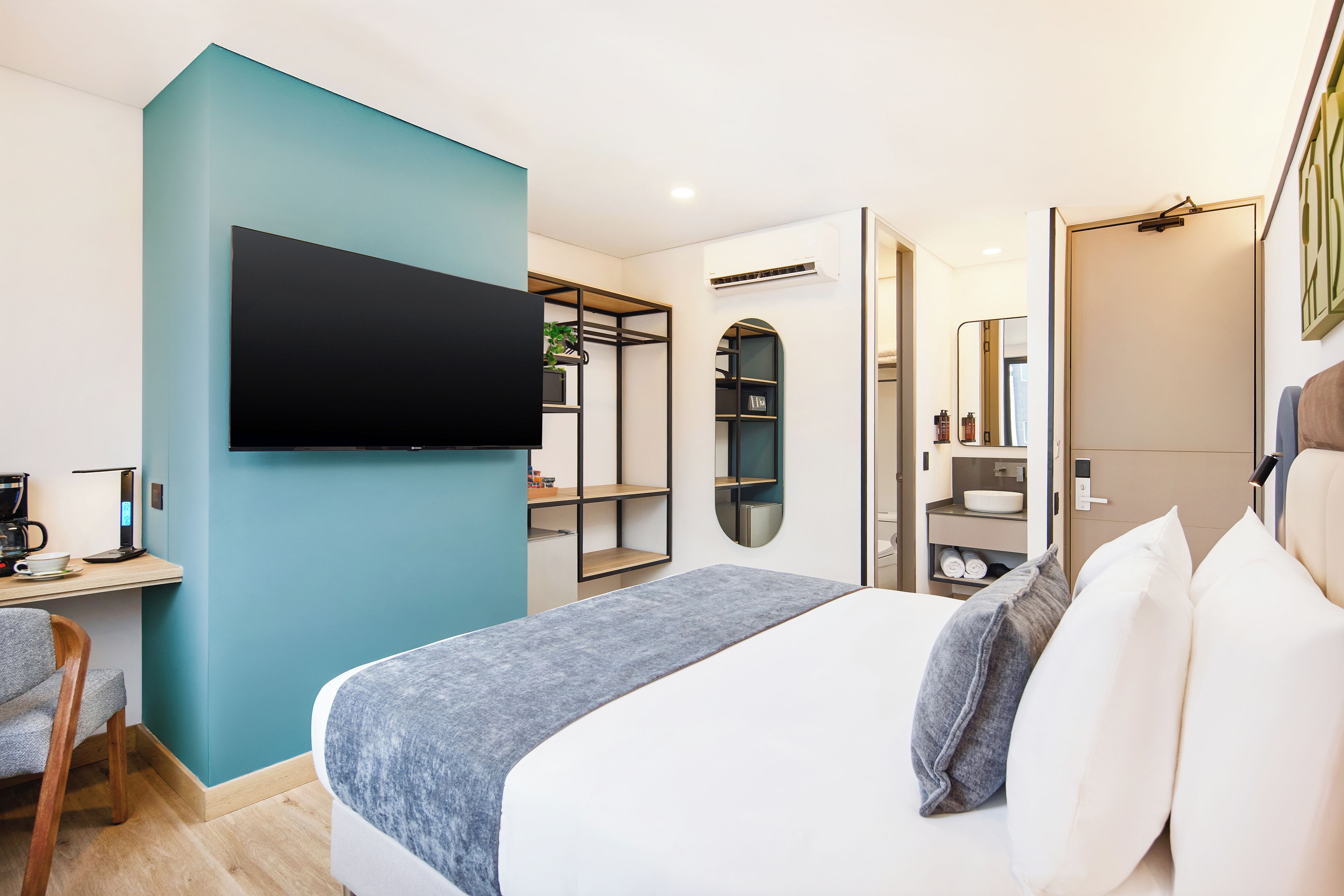 A modern hotel room with a bed, a desk with a coffee maker, a TV mounted on the wall, and a wardrobe with shelves.