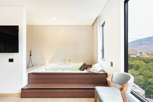 A modern bathroom with a freestanding bathtub, a wooden bench, and a view of a cityscape through a large window.