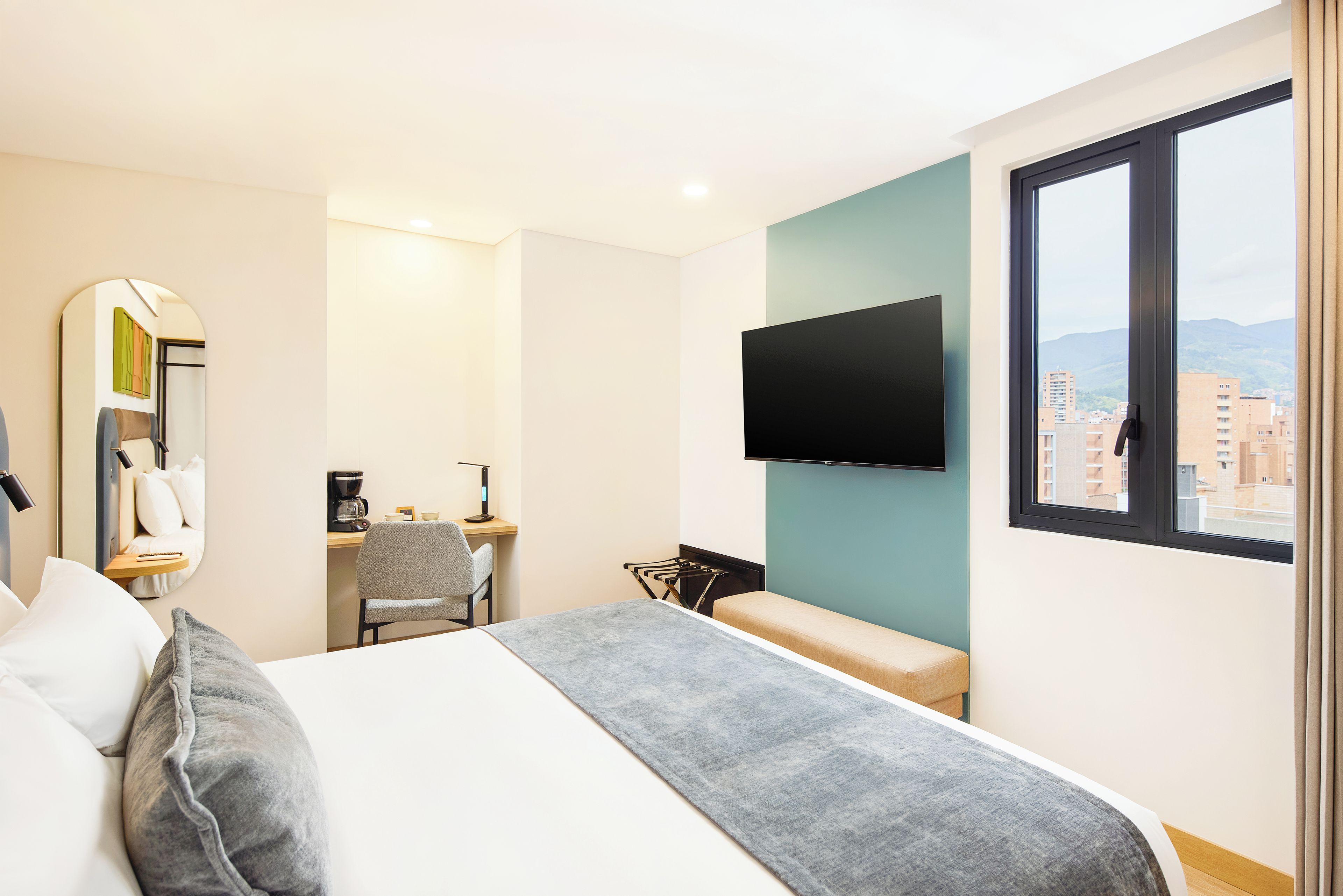 A hotel room with a bed, a desk, a chair, a TV, and a view of the city.