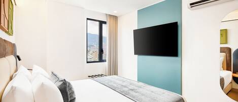 A hotel room with a bed, a mounted TV, a window with a city view, and a wall-mounted air conditioning unit.