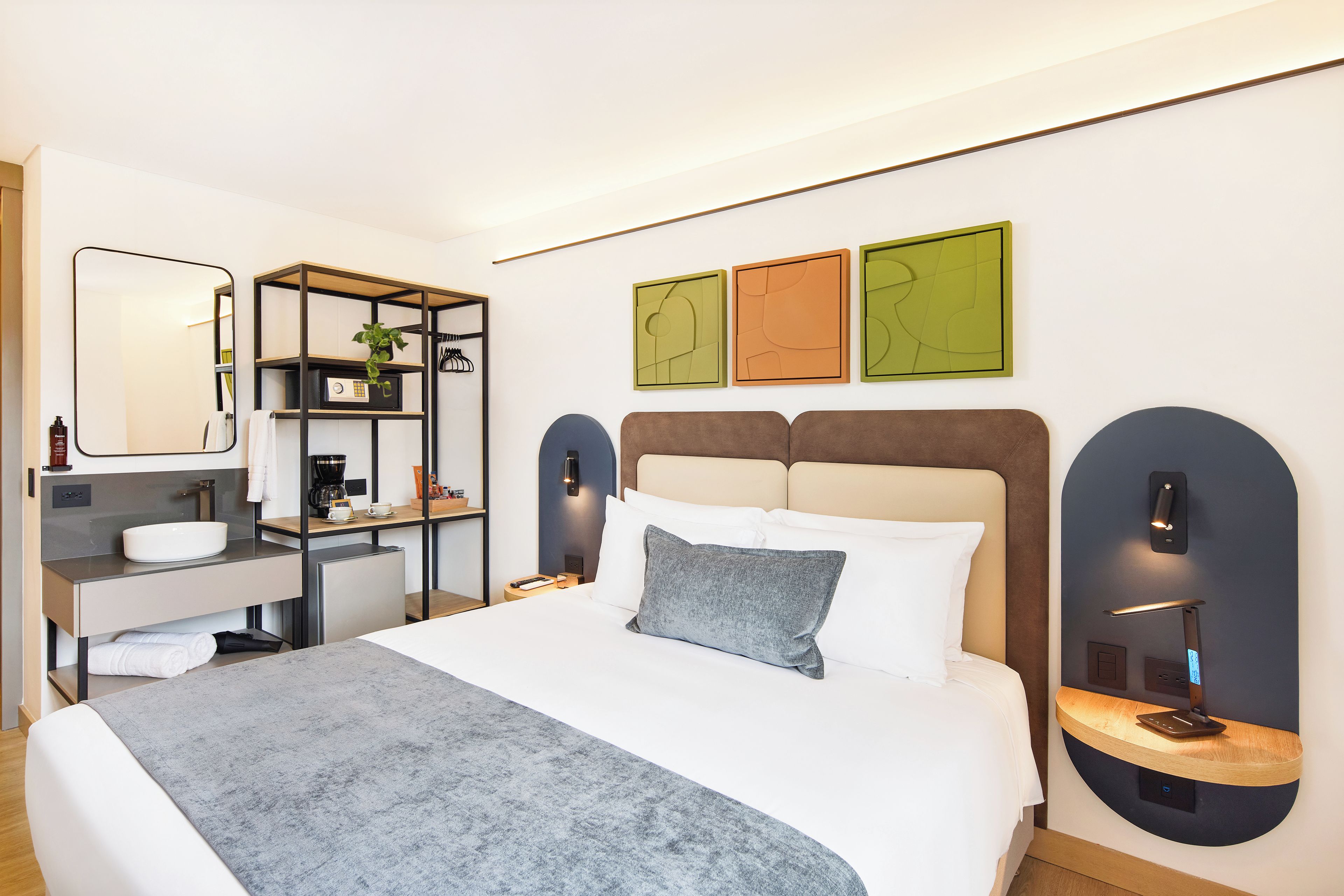 A modern hotel room with a bed, bedside tables, a desk, a mirror, and artwork on the wall.