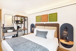 Quarto de hotel moderno com cama, mesas de cabeceira, escrivaninha, espelho e obras de arte na parede.